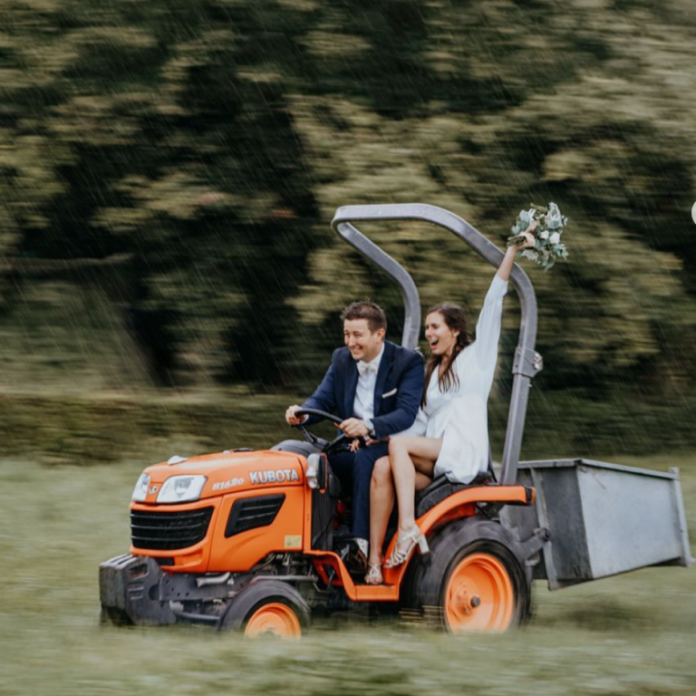Un couple joyeux sur un tracteur, la mariée levant un bouquet, en pleine nature.